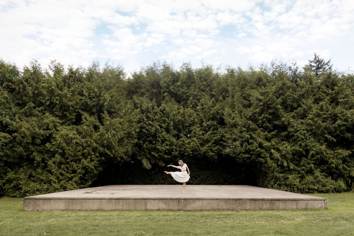 Ballerina in black tutu and pink top poses on colorful urban wall surrounded by green trees.