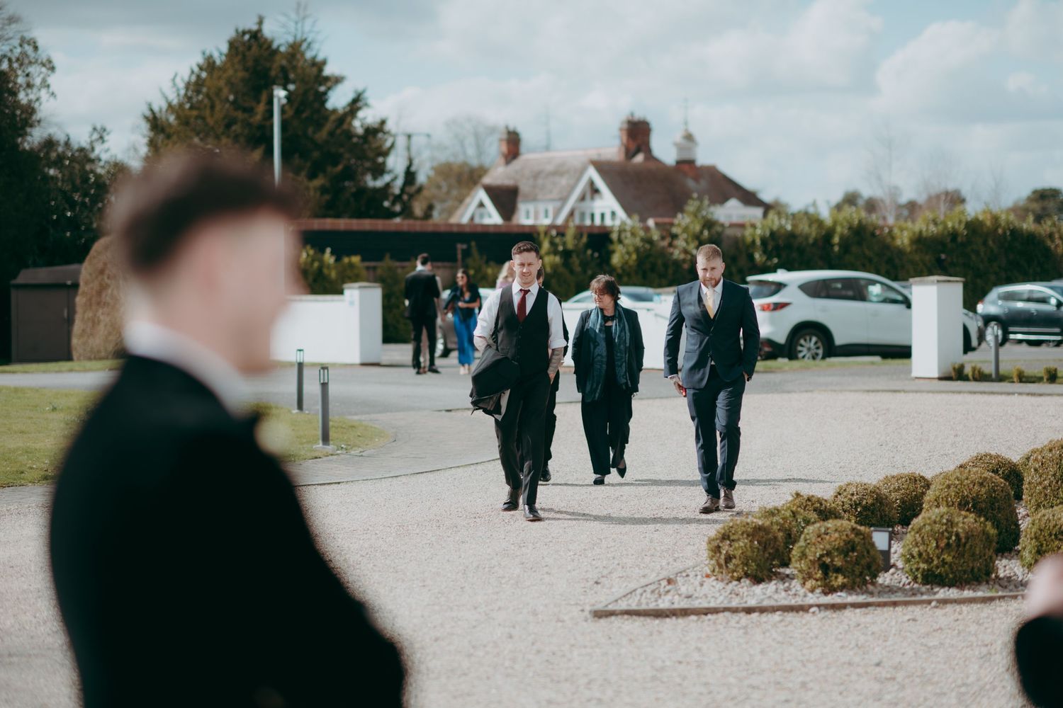 Groomsmen in black suits walking along a gravel path toward the wedding venue on a bright day.
