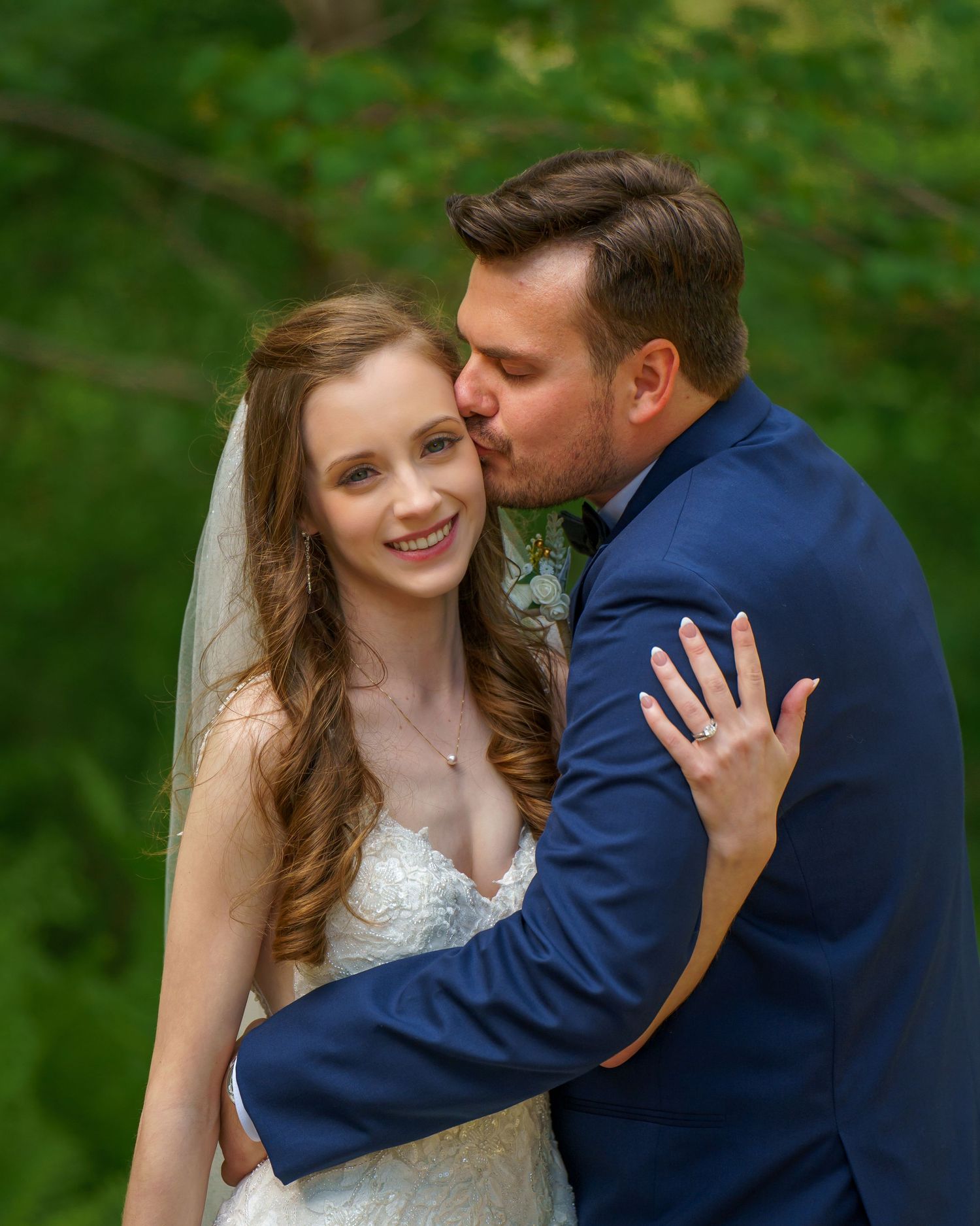 A groom in a blue suit kisses his smiling bride on the cheek outdoors, with lush green foliage in the background.