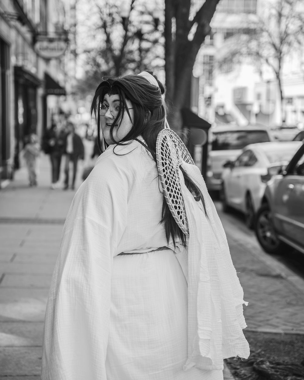 Black and white street photo of a woman in a white robe with decorative back detail, standing on an urban sidewalk.