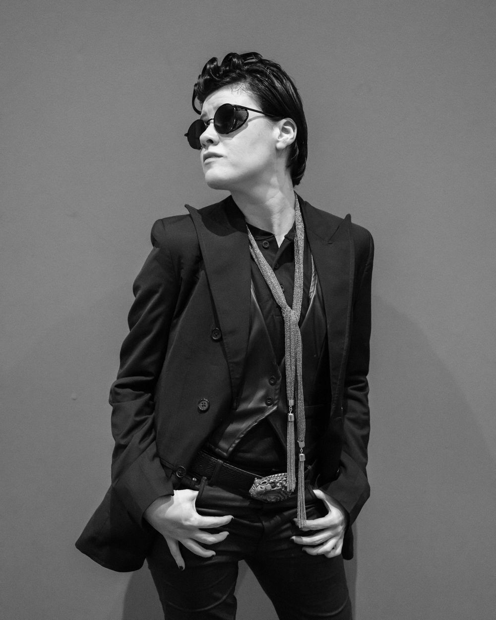 Black and white photo of a stylish person in a blazer and sunglasses posing confidently against a neutral background.
