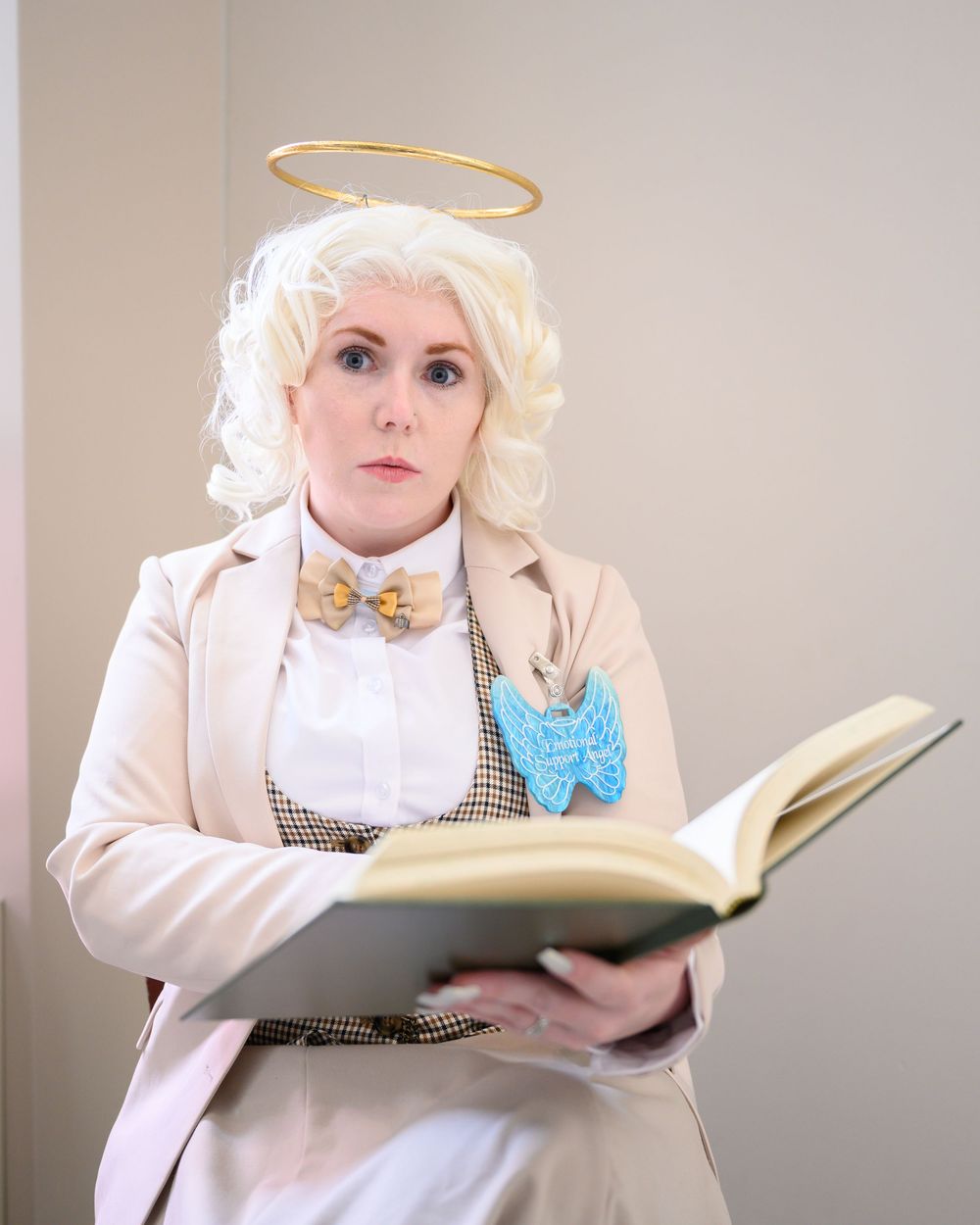Woman cosplaying as an angel with halo, blonde wig, bow tie, and blue badge, holding an open book.