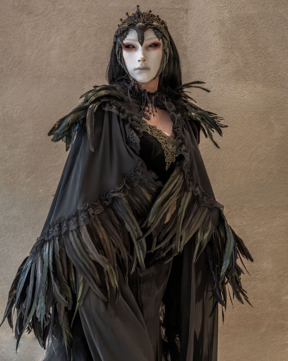 Person in dark gothic costume with feathered cape, white mask, red eyes, and black crown.