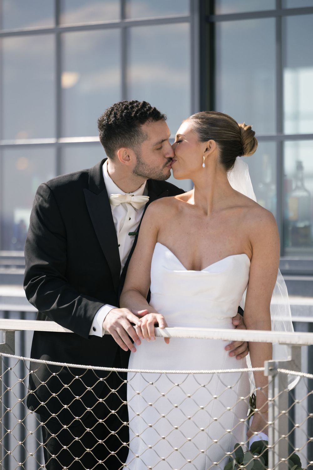 Bride and groom share a romantic kiss on a balcony, dressed in a white strapless gown and black tuxedo.