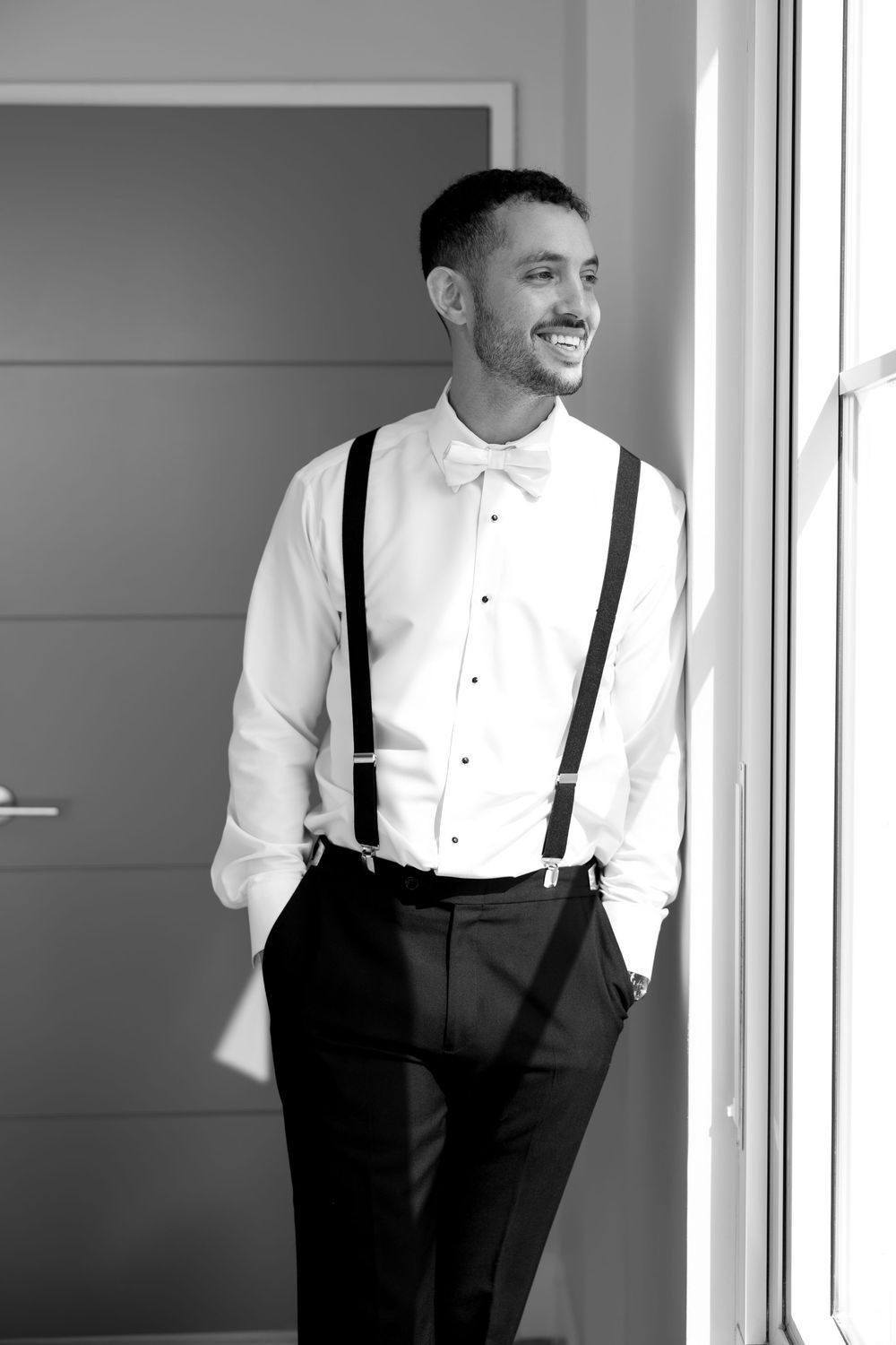 Smiling groom in white dress shirt, bow tie, and suspenders stands by window in black and white wedding photo.