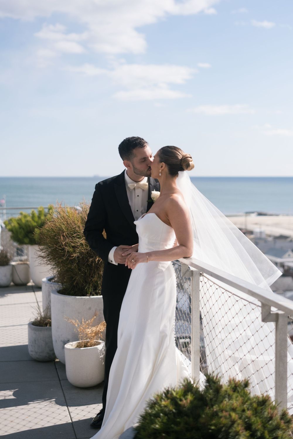 Bride and groom share a romantic kiss on an oceanfront terrace, her veil flowing in the breeze.
