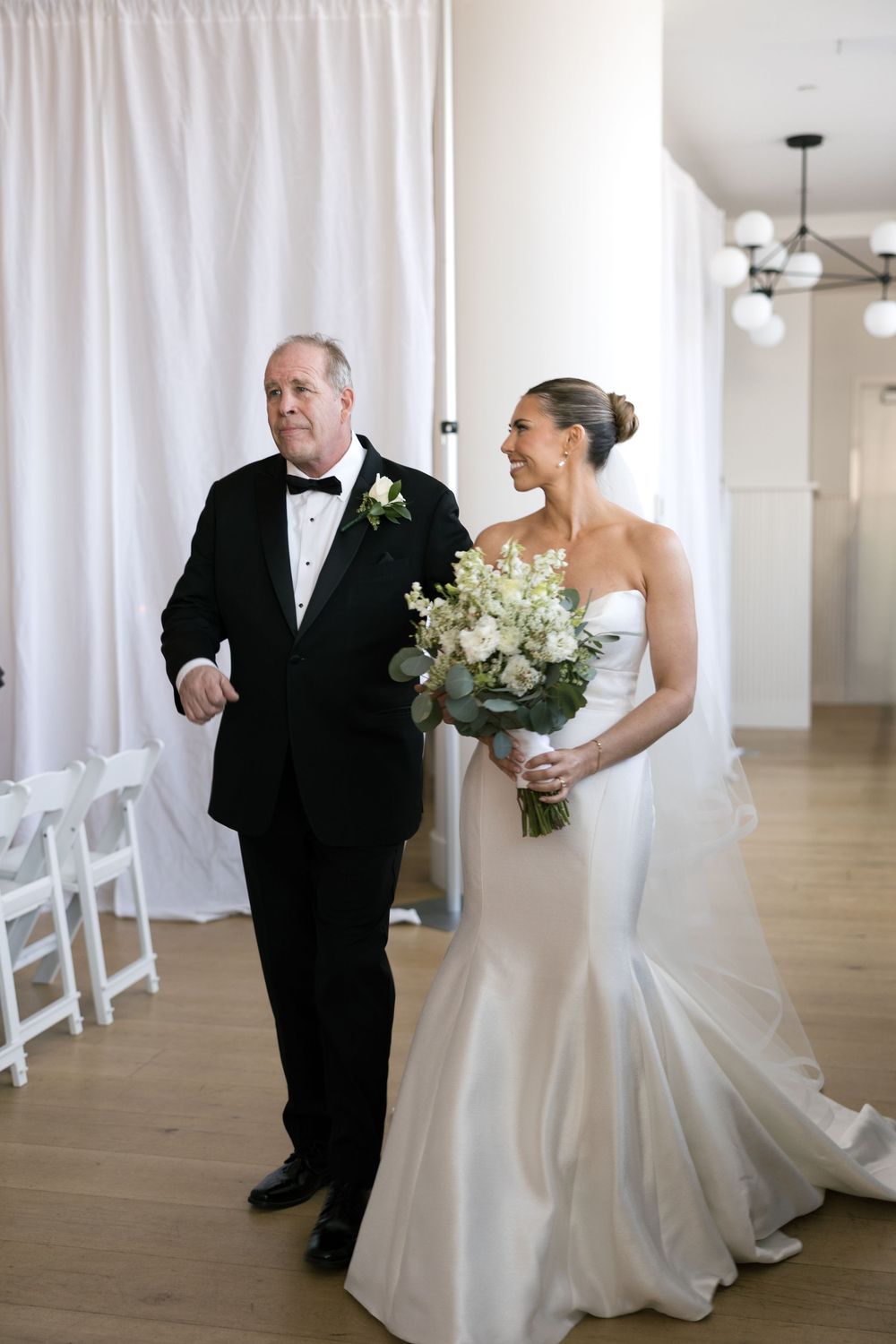 Bride in strapless satin mermaid gown holding white bouquet walks down aisle with escort in black tuxedo.