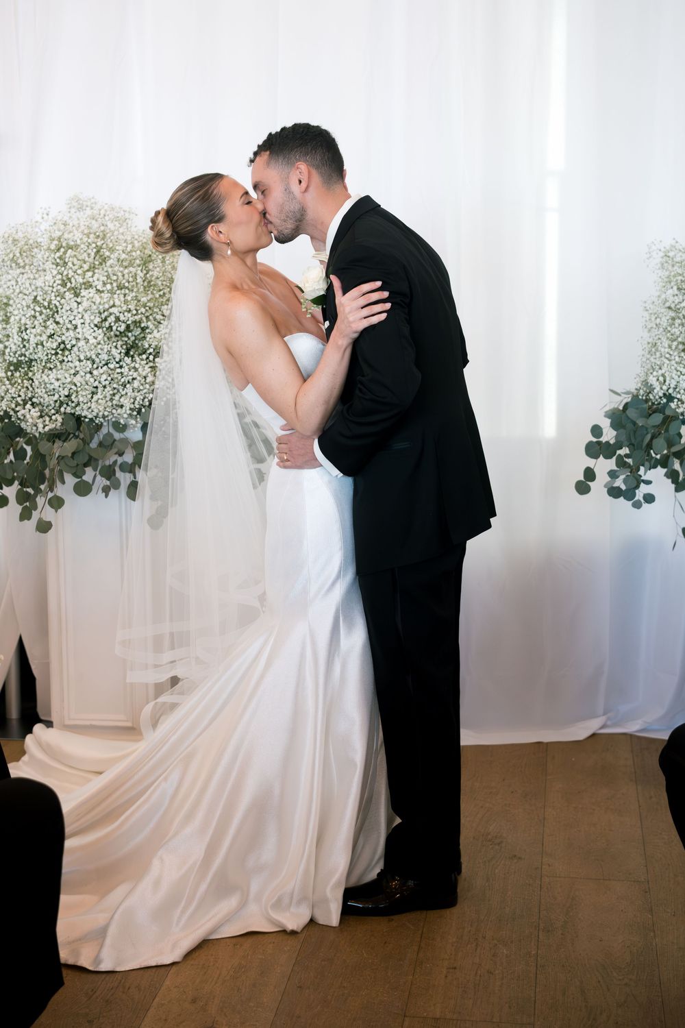 Bride and groom share their first kiss at the altar, surrounded by baby's breath floral arrangements.