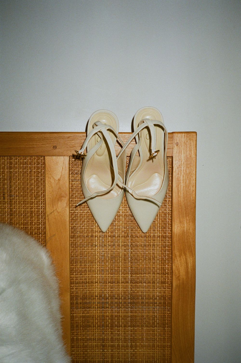 Cream patent leather pointed-toe mules with ankle straps displayed on a woven rattan headboard.