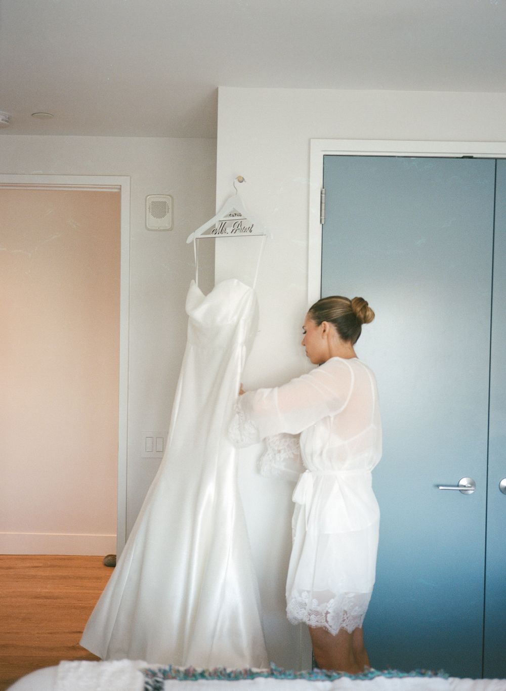 Bride in white lace robe admires her elegant wedding dress hanging on a personalized hanger indoors.