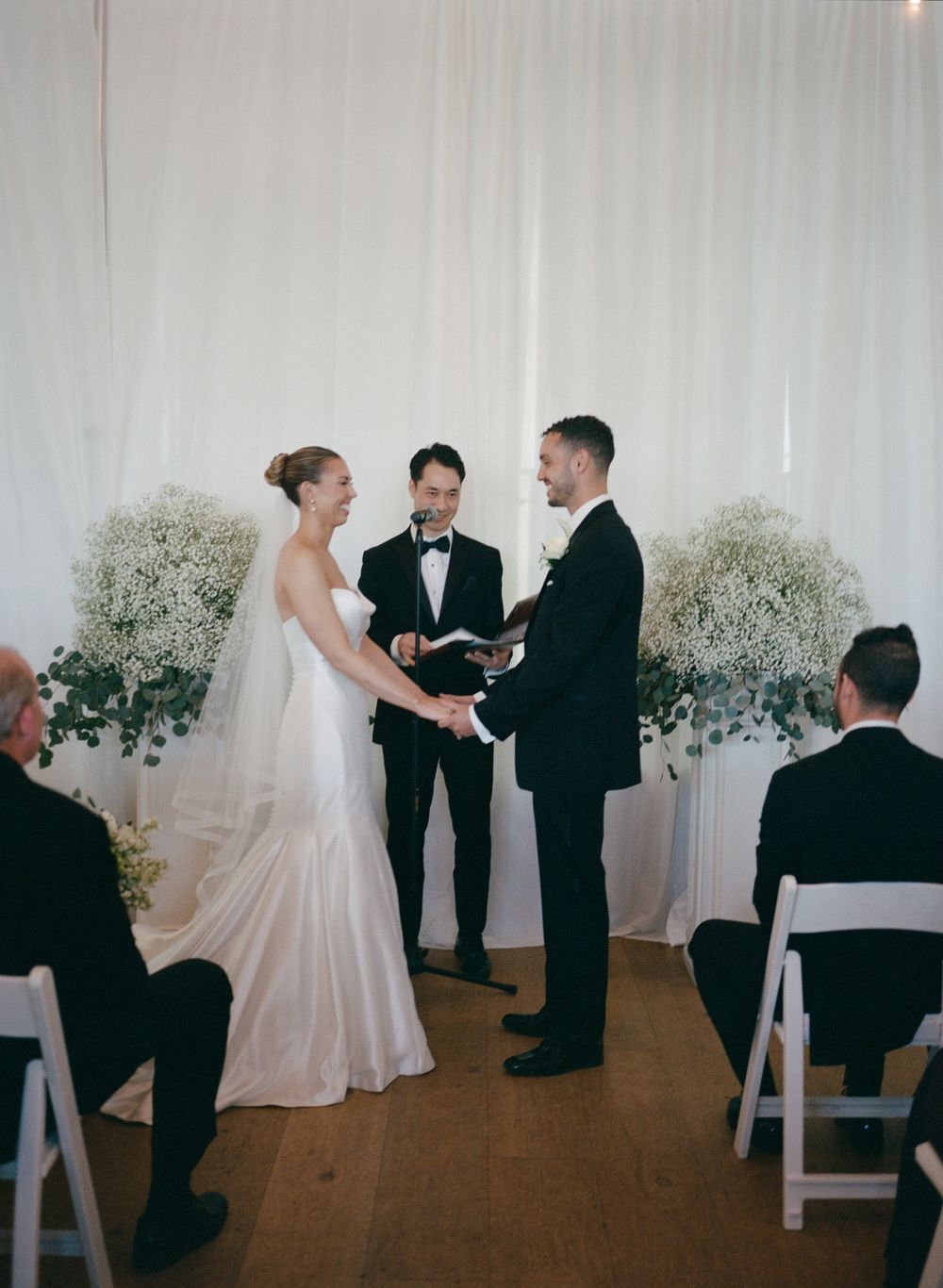 Bride and groom hold hands at elegant indoor wedding ceremony with white draping and baby's breath floral arrangements.