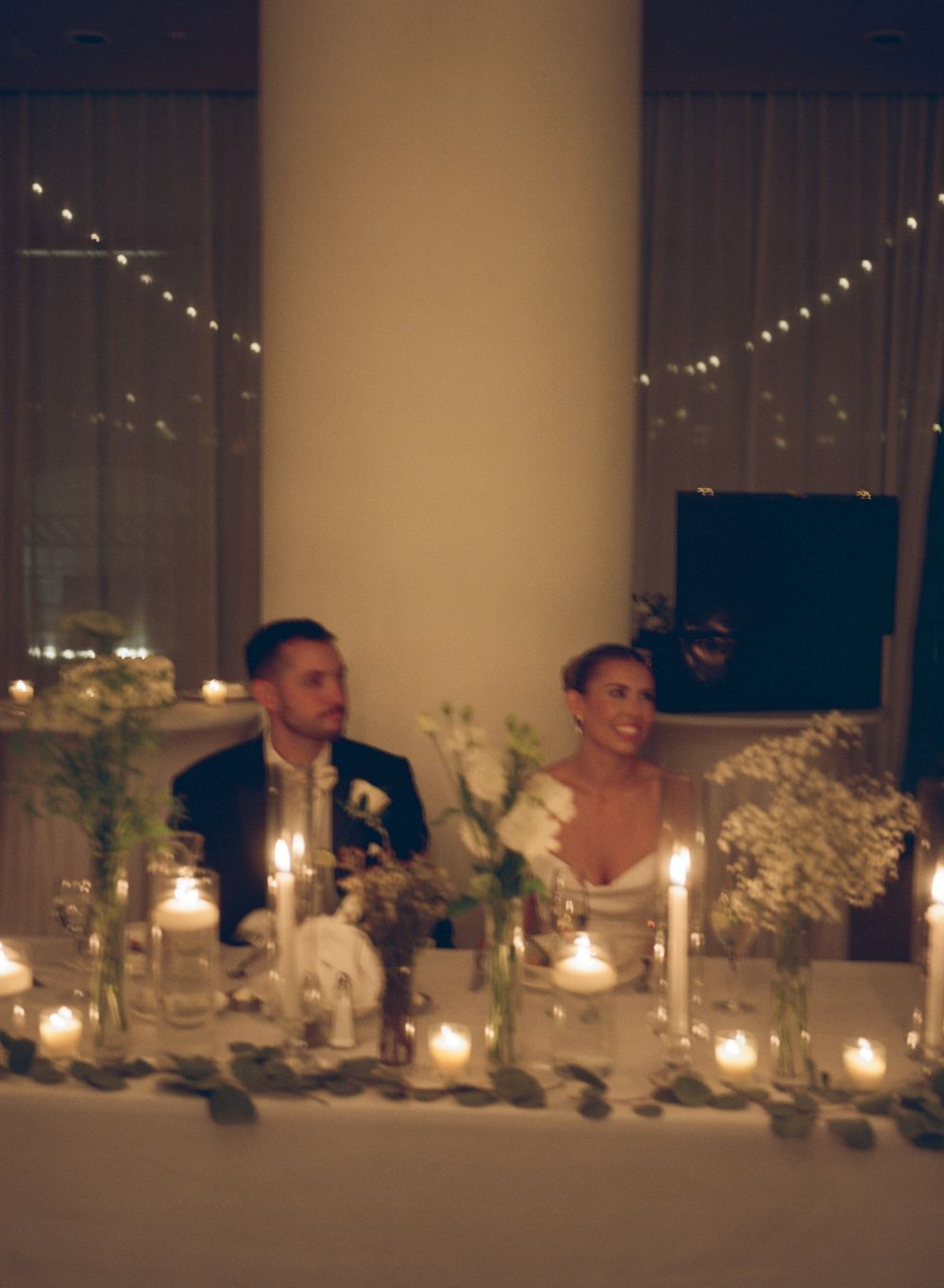 Bride and groom seated at candlelit wedding reception sweetheart table with floral arrangements and fairy lights.
