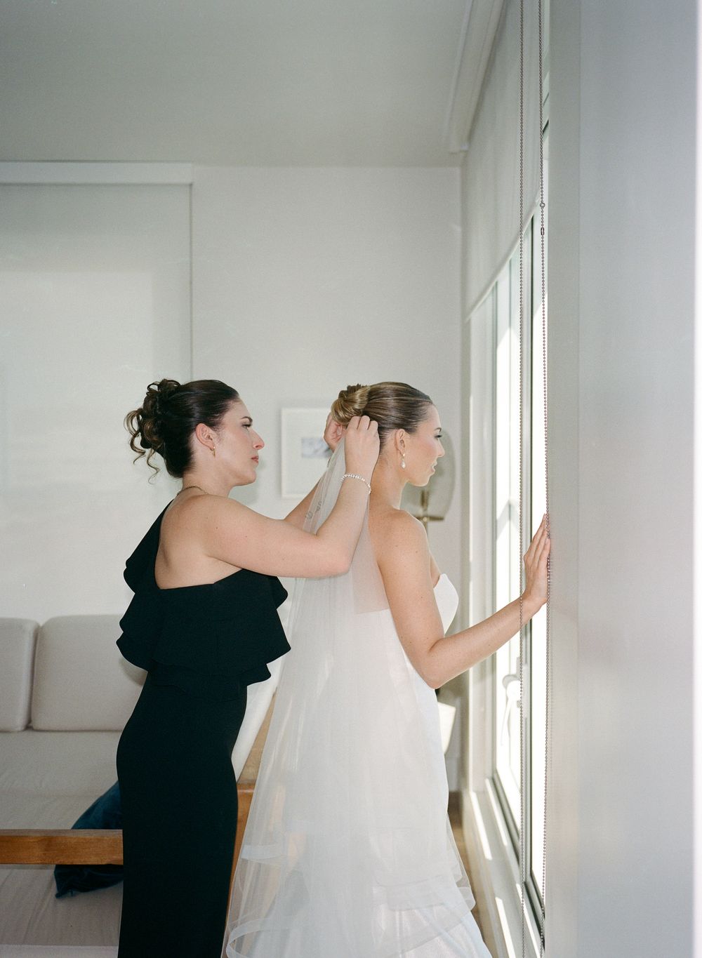 Bridesmaid in black dress adjusting bride's veil near window during wedding preparations.