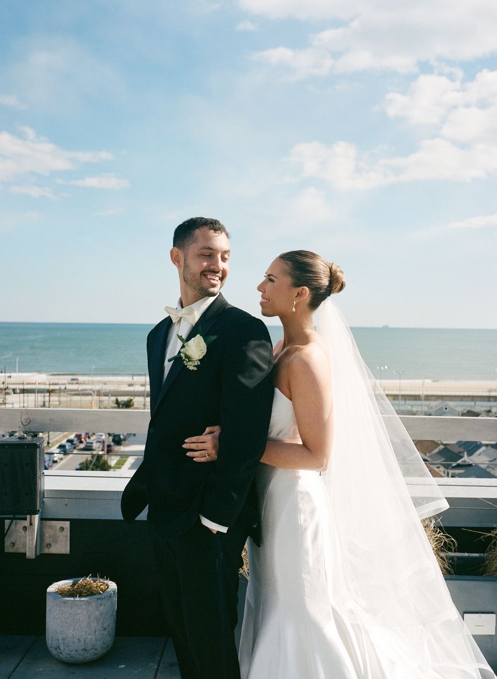 Smiling bride and groom pose on a rooftop terrace with a stunning ocean and boardwalk backdrop.