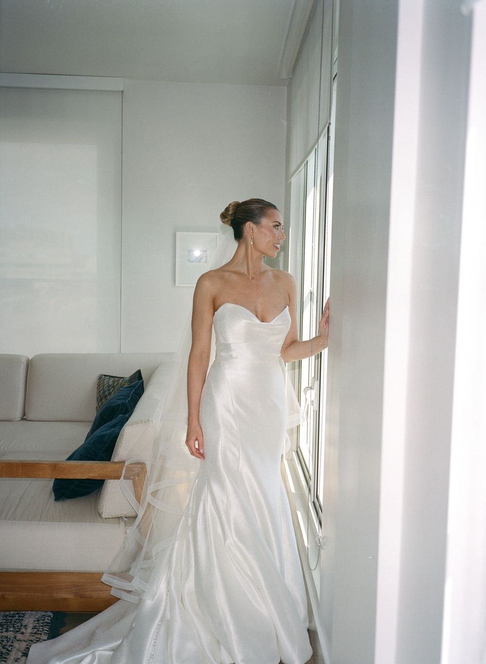 Bride in elegant strapless satin mermaid wedding gown with train stands by window in natural light.