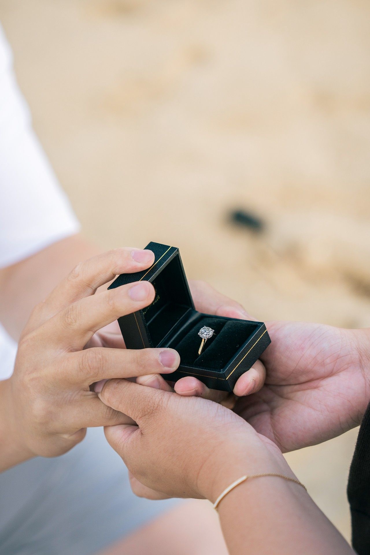 Person holding open a black ring box with a diamond engagement ring on a sandy beach during a marriage proposal.