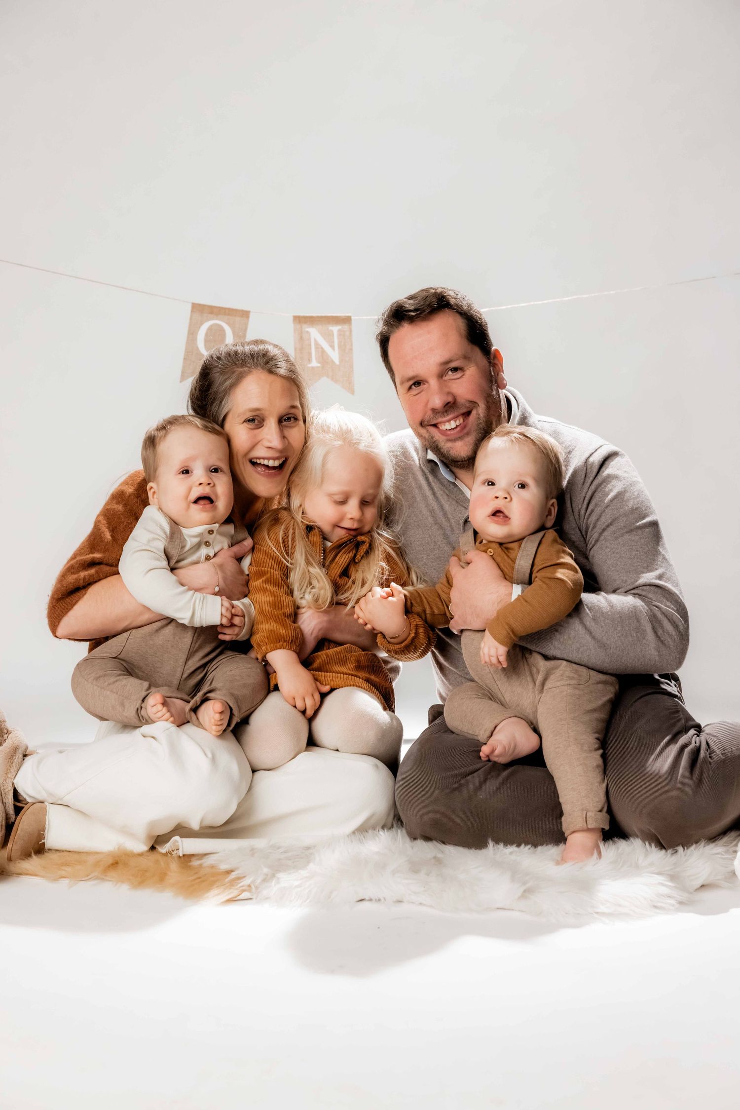 Happy family with two parents and three young children sitting together on a white fur rug in a bright studio photo.