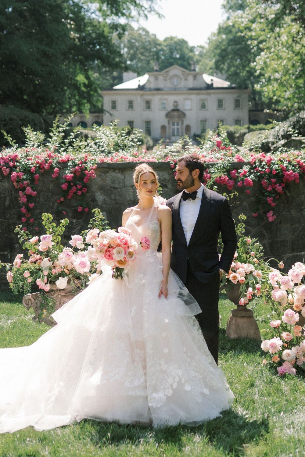 Bride in tiered lace gown holds pink bouquet beside groom in tuxedo, posing before floral arch and elegant manor.