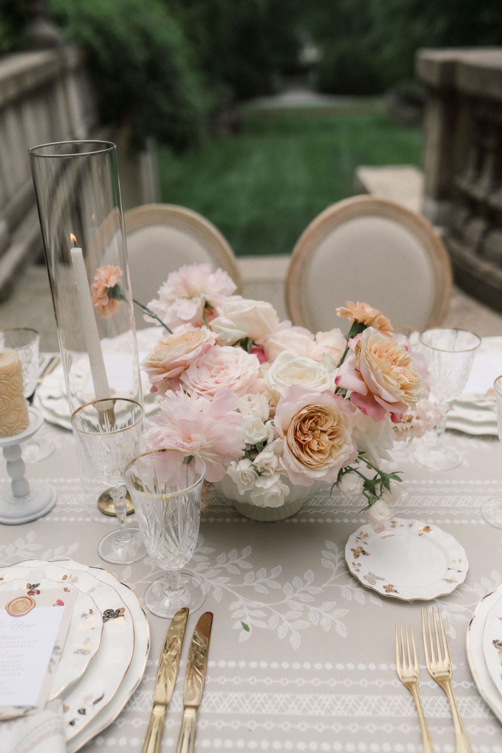 Elegant wedding table setting with blush pink roses, gold cutlery, crystal glasses, and a candle centerpiece on a lace tablecloth.