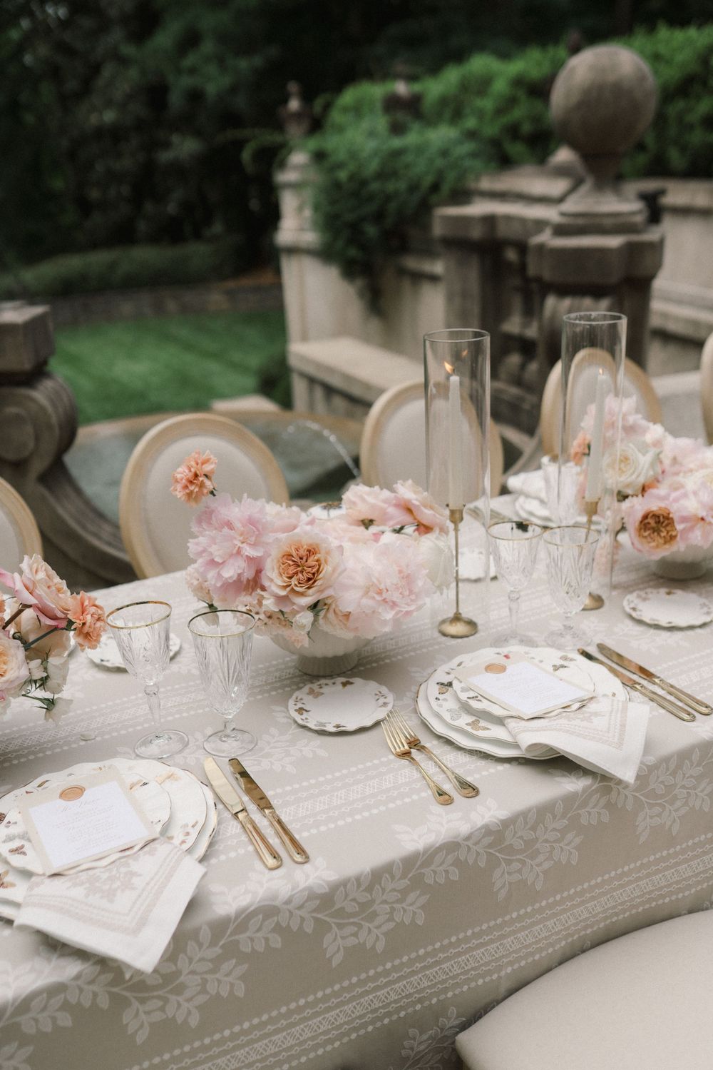 Elegant outdoor wedding tablescape with pink peonies, gold cutlery, crystal glasses, and floral linen on a garden terrace.