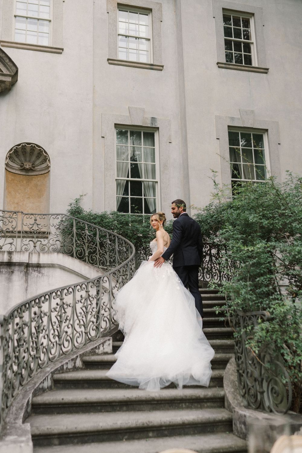 Bride and groom embrace on ornate spiral staircase outside elegant stone mansion surrounded by lush greenery.