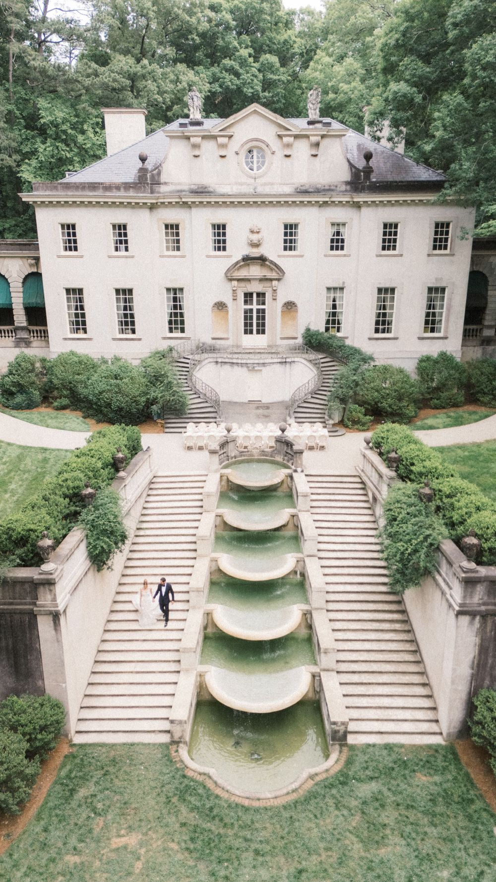 Aerial view of an elegant Georgian mansion with cascading fountain steps and lush greenery, featuring a couple walking the grounds.