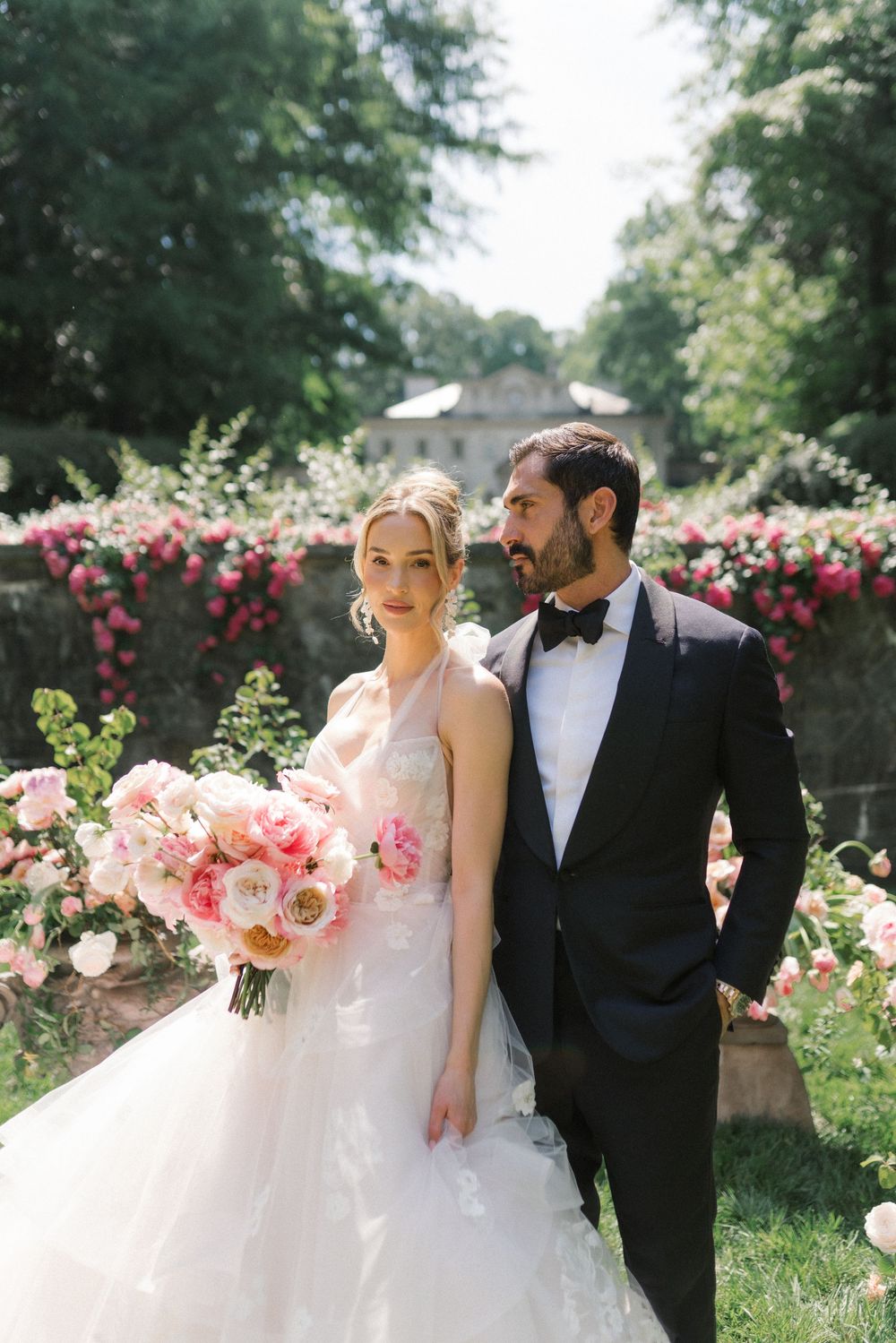 Bride and groom pose in lush garden with pink roses, she holds a pink bouquet in a white gown, he wears a black tuxedo.