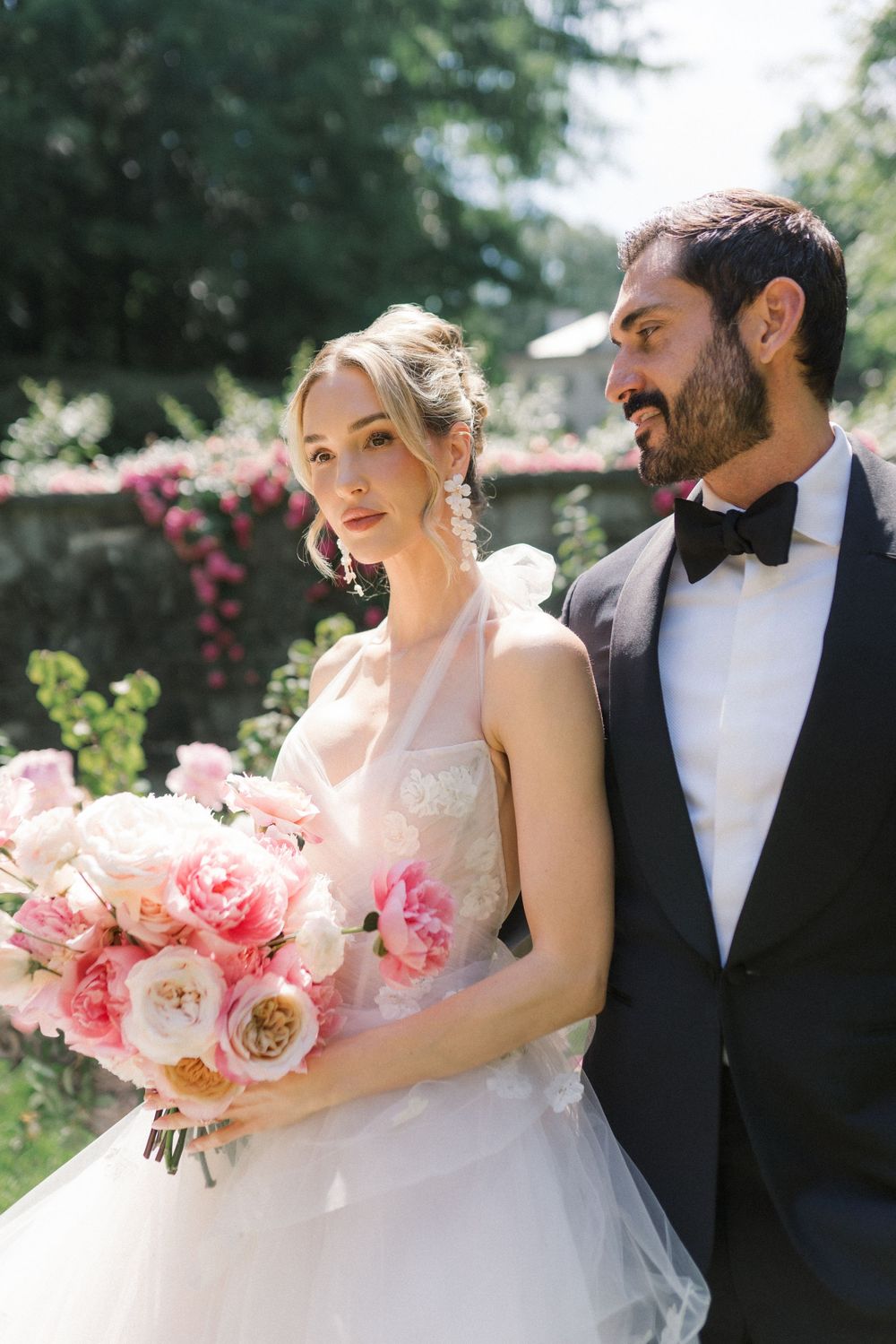 Bride holding pink peony bouquet in white gown stands with groom in black tuxedo in a sunlit garden.