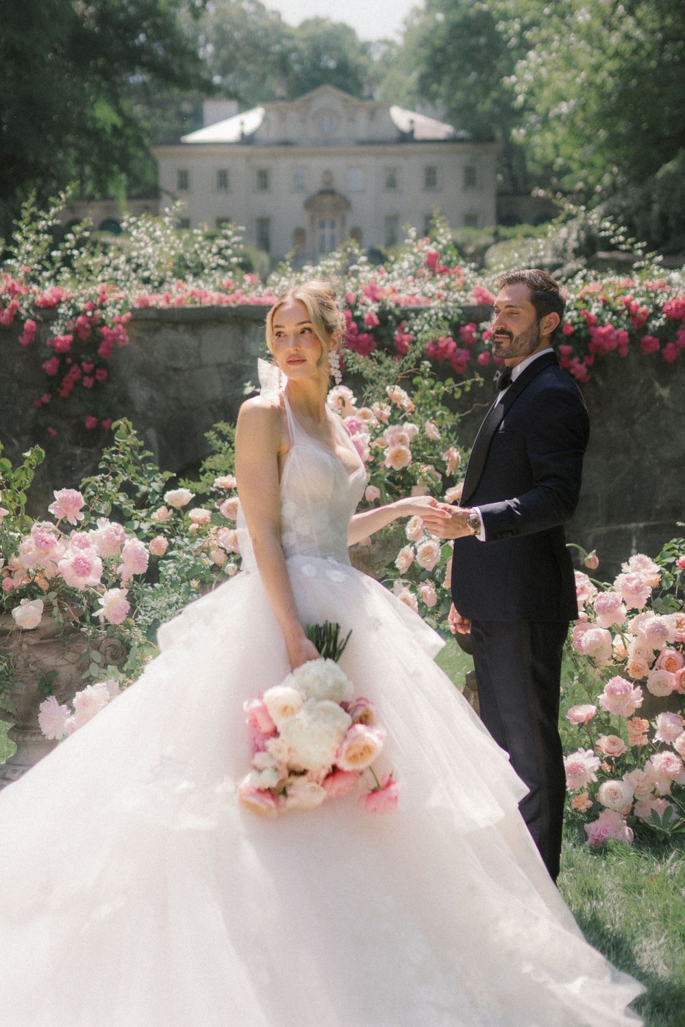 Bride in a layered tulle gown holds a pink bouquet beside a tuxedo-clad groom, surrounded by lush rose gardens.