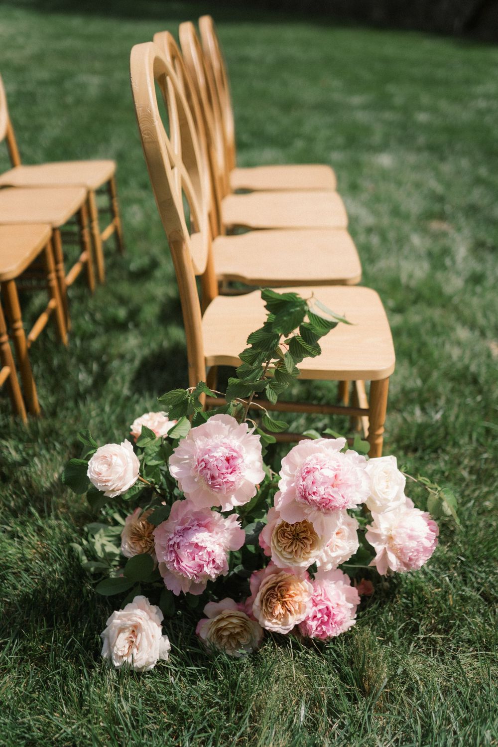 Pink peony bouquet on grass beside gold chiavari chairs at an elegant outdoor wedding ceremony.