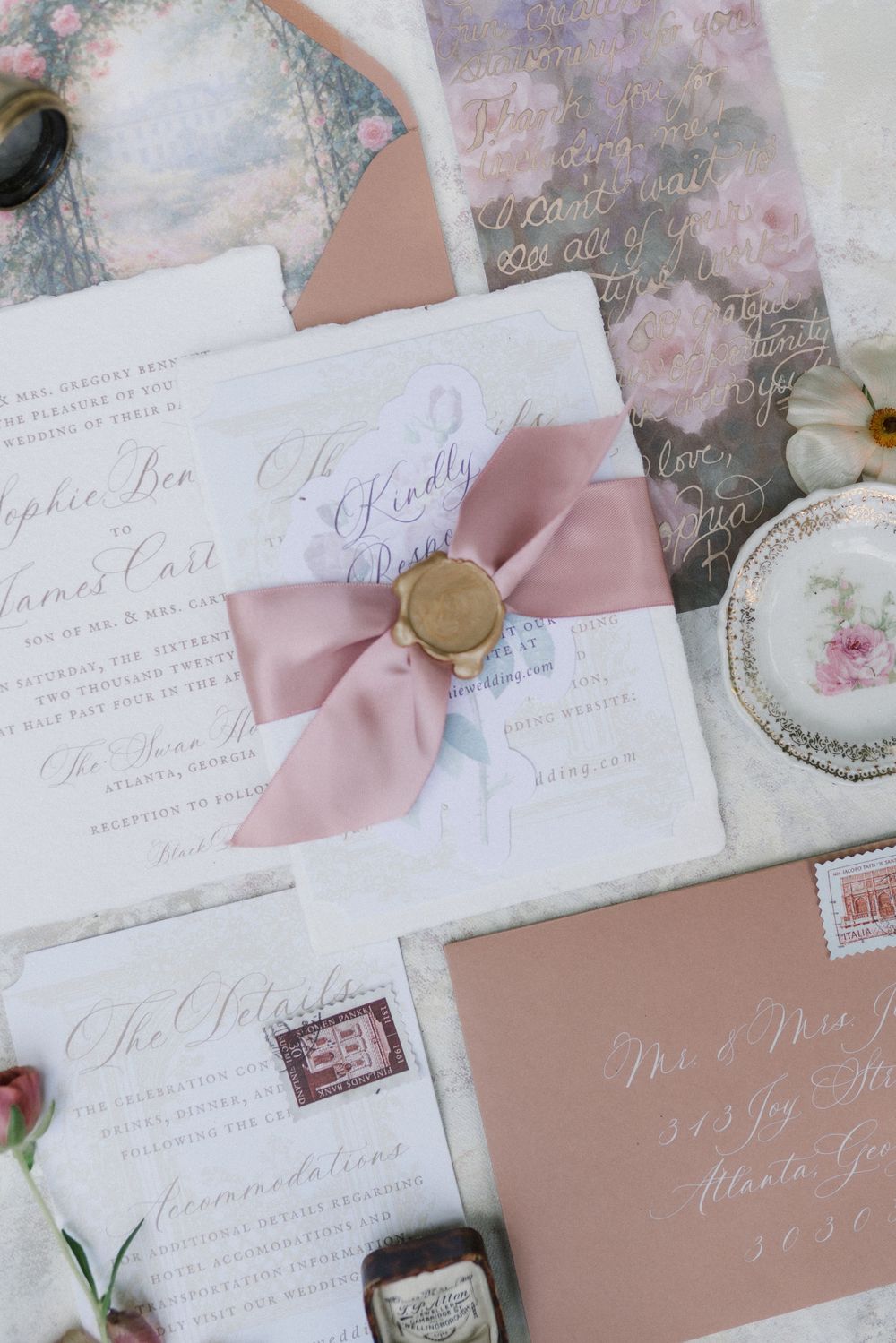 Elegant wedding invitation suite with dusty rose ribbon, gold wax seal, calligraphy, and floral accents on soft white stationery.