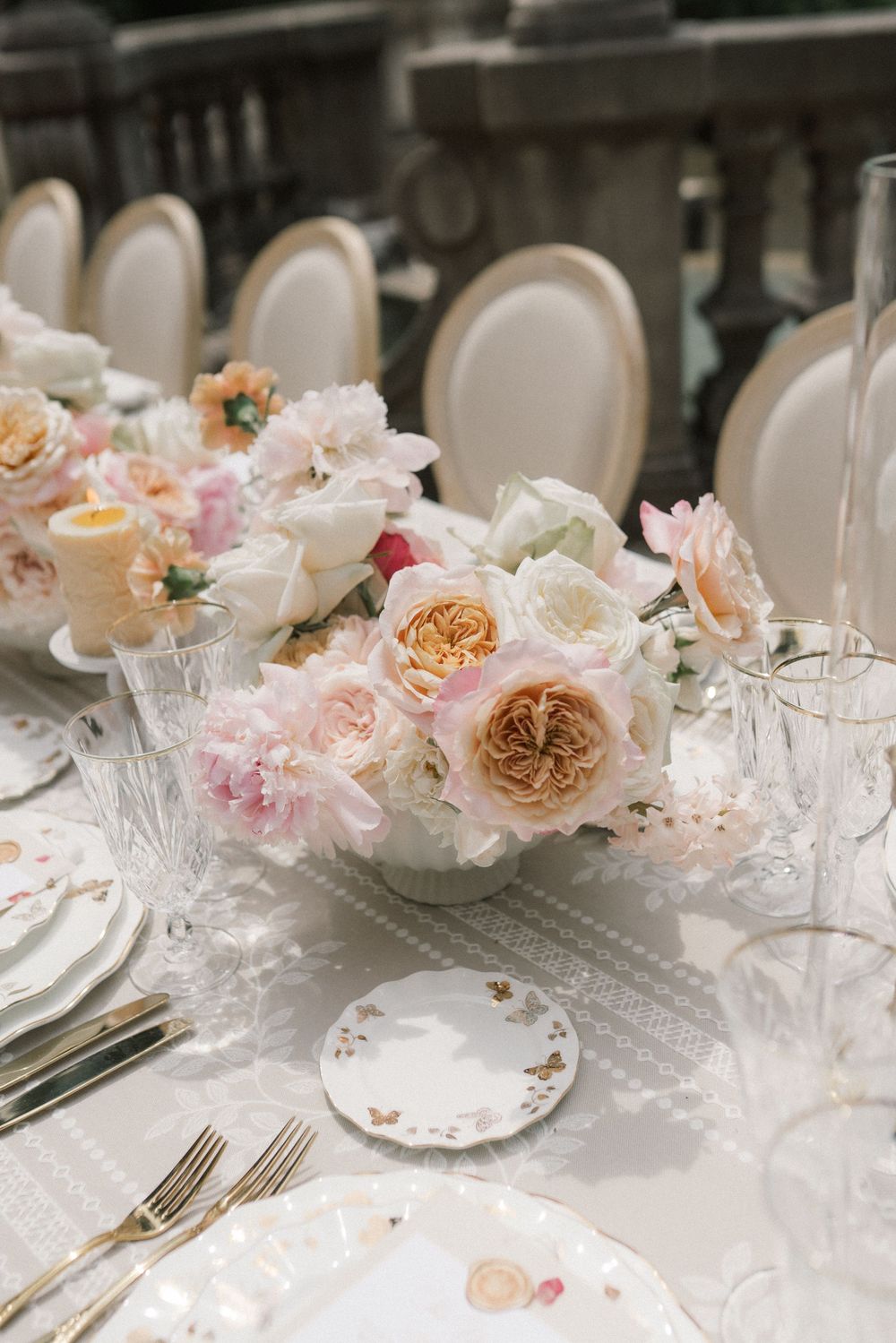 Elegant wedding reception table with pink and peach floral centerpiece, crystal glasses, gold cutlery, and floral china plates.