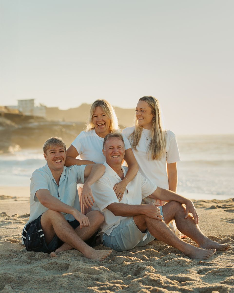 Happy family of four sitting together on sandy beach at golden hour, smiling and wearing white outfits bronte beach family photoshoot Sydney