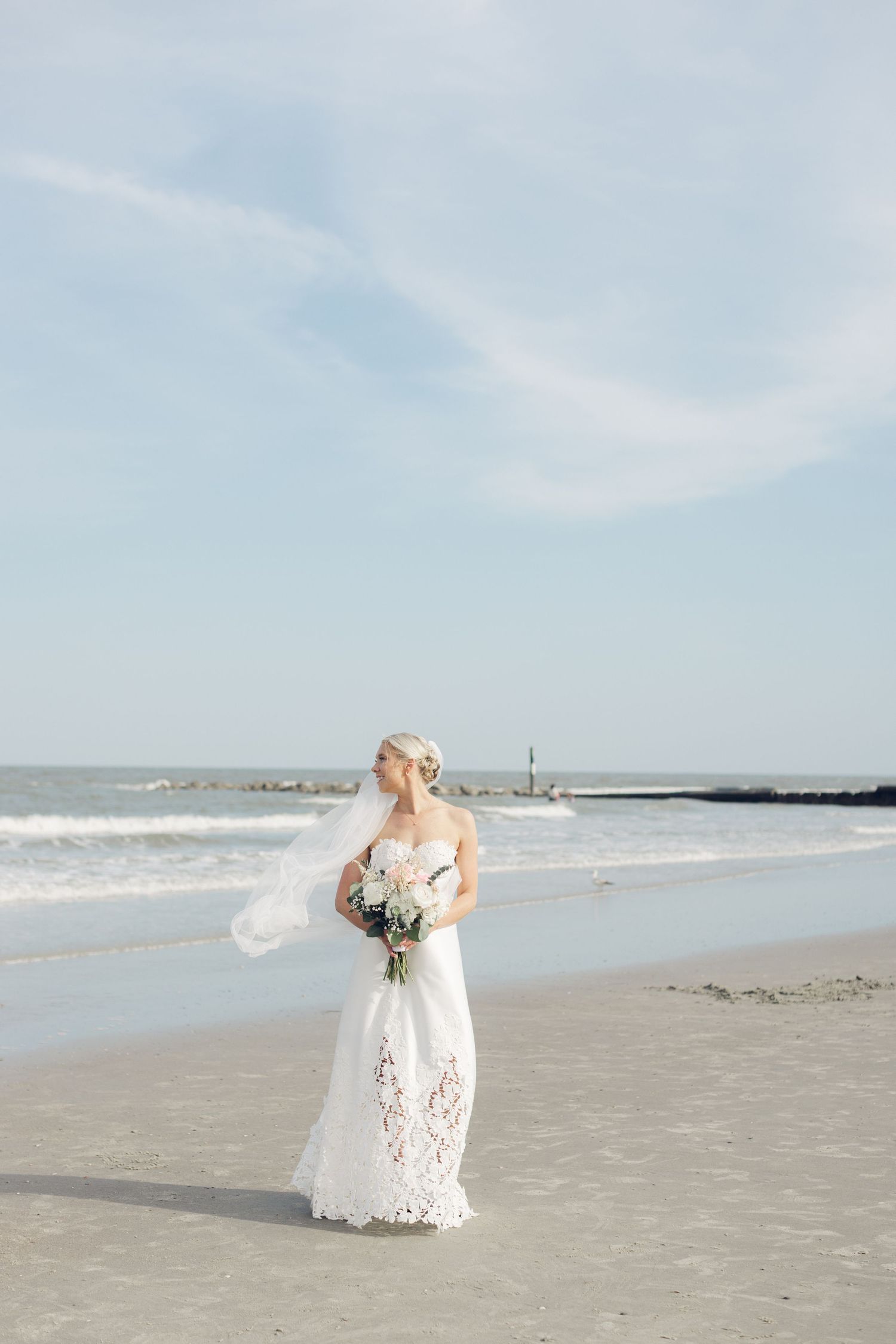 Couple shares romantic dip kiss on sandy beach with wedding party celebrating behind them near a lighthouse.