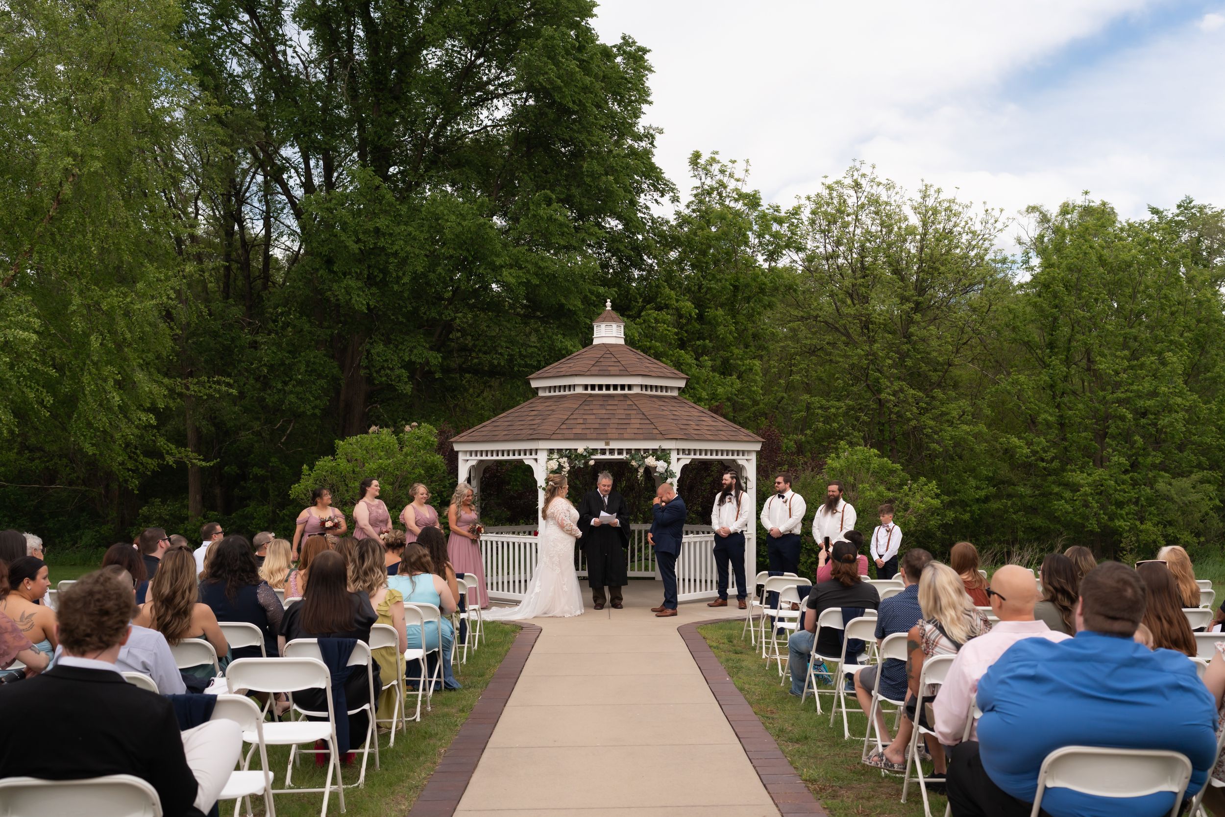 Outdoor wedding ceremony at a gazebo surrounded by lush green trees with guests seated in white chairs.