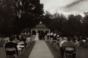 Black and white photo of outdoor wedding ceremony at gazebo with guests seated in white chairs.