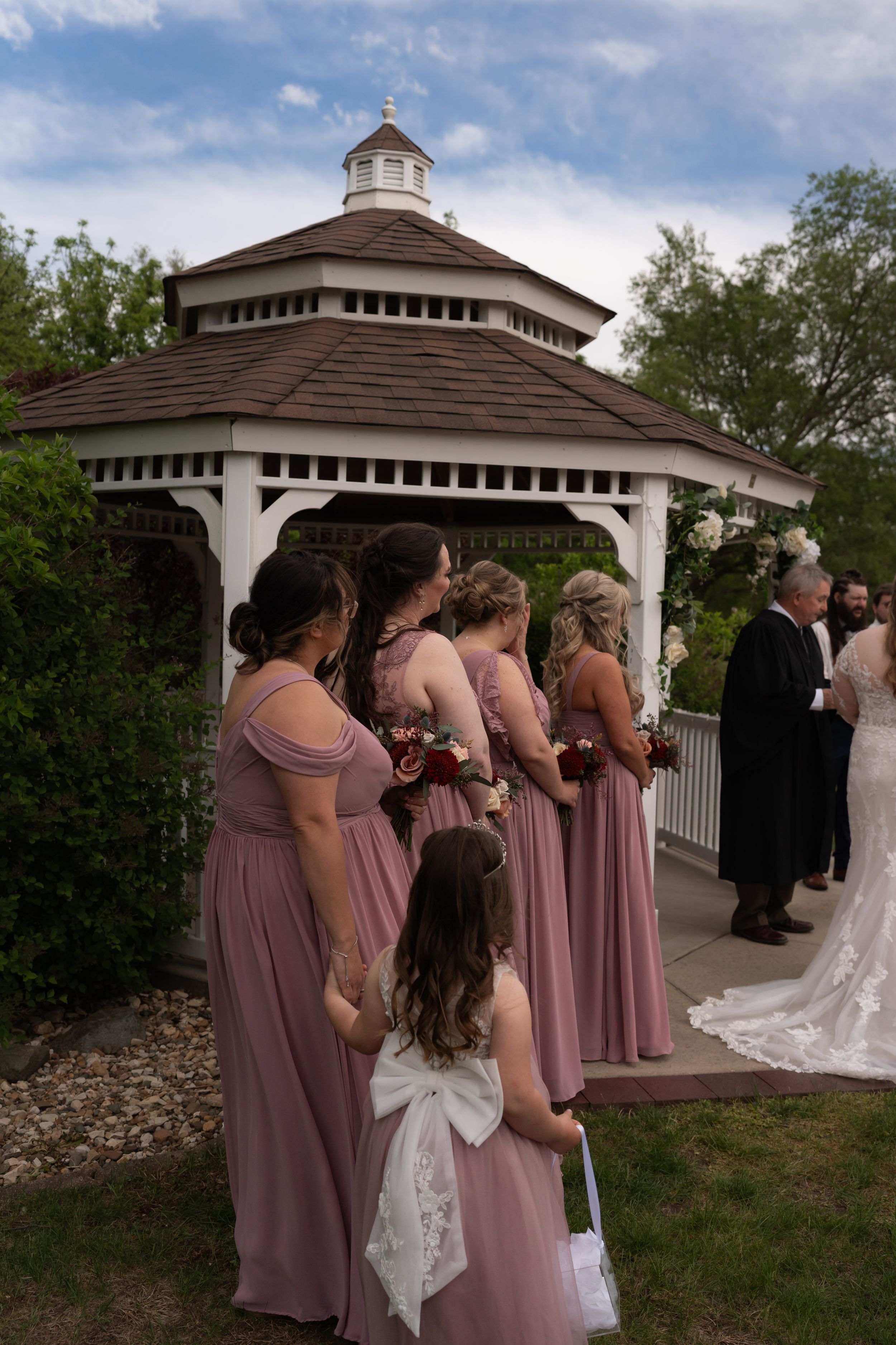 Bridesmaids in dusty rose gowns stand near a white gazebo during an outdoor wedding ceremony.