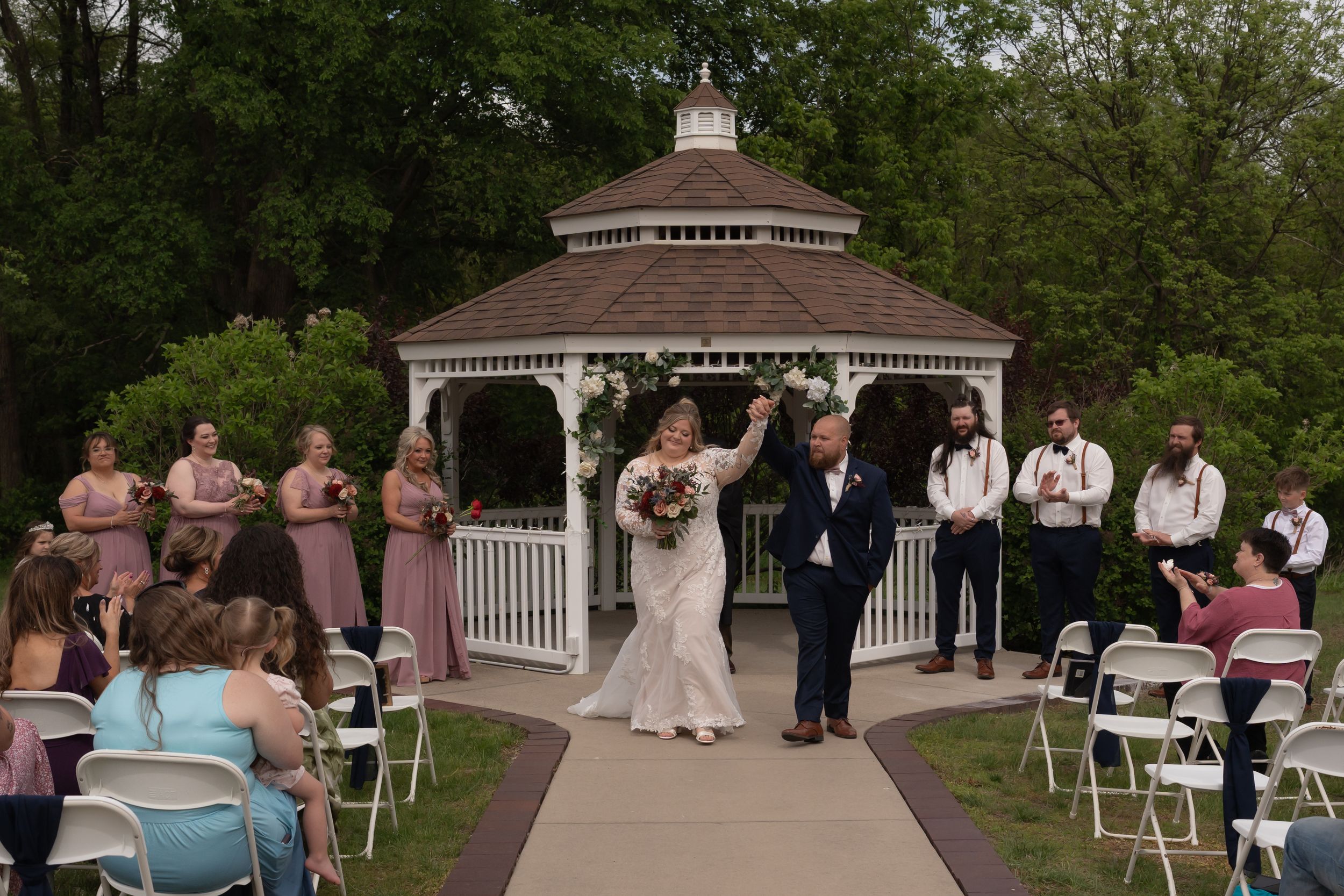 Bride and groom celebrate at outdoor wedding ceremony near white gazebo, surrounded by bridesmaids and guests in garden setting.