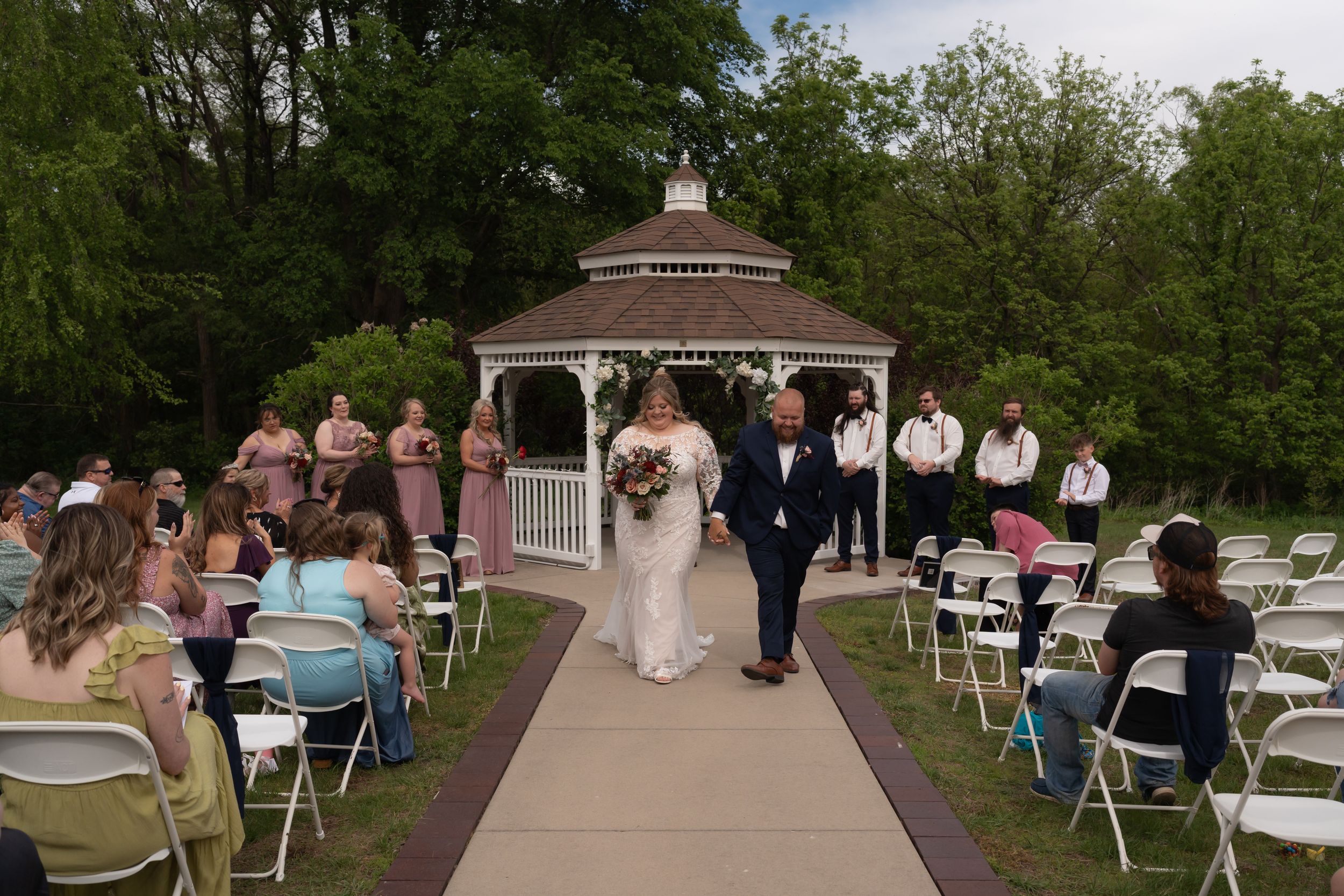 Bride and groom walk down outdoor aisle after wedding ceremony at a gazebo venue surrounded by guests.