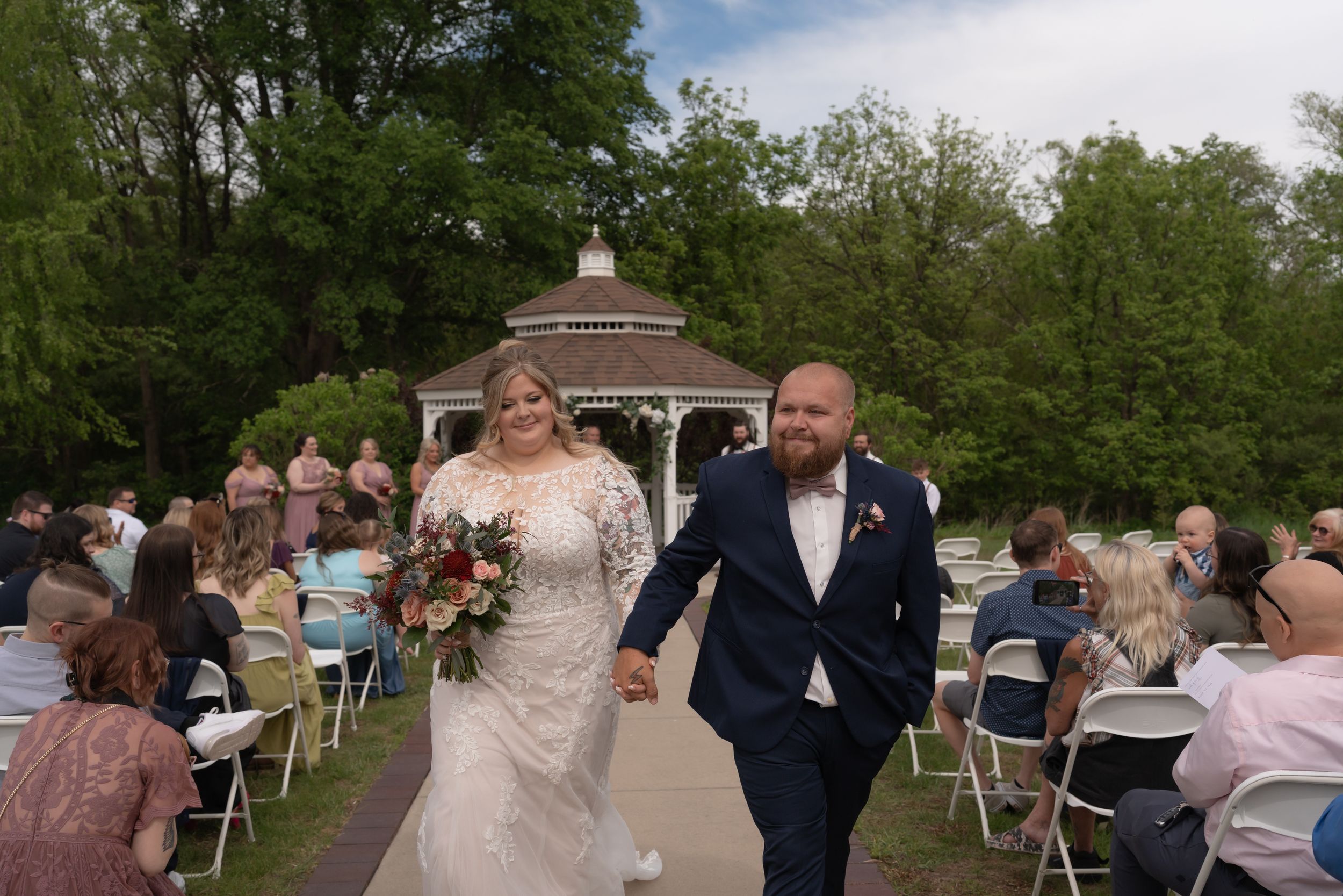 Bride and groom walk hand-in-hand down outdoor aisle after wedding ceremony near gazebo surrounded by seated guests.