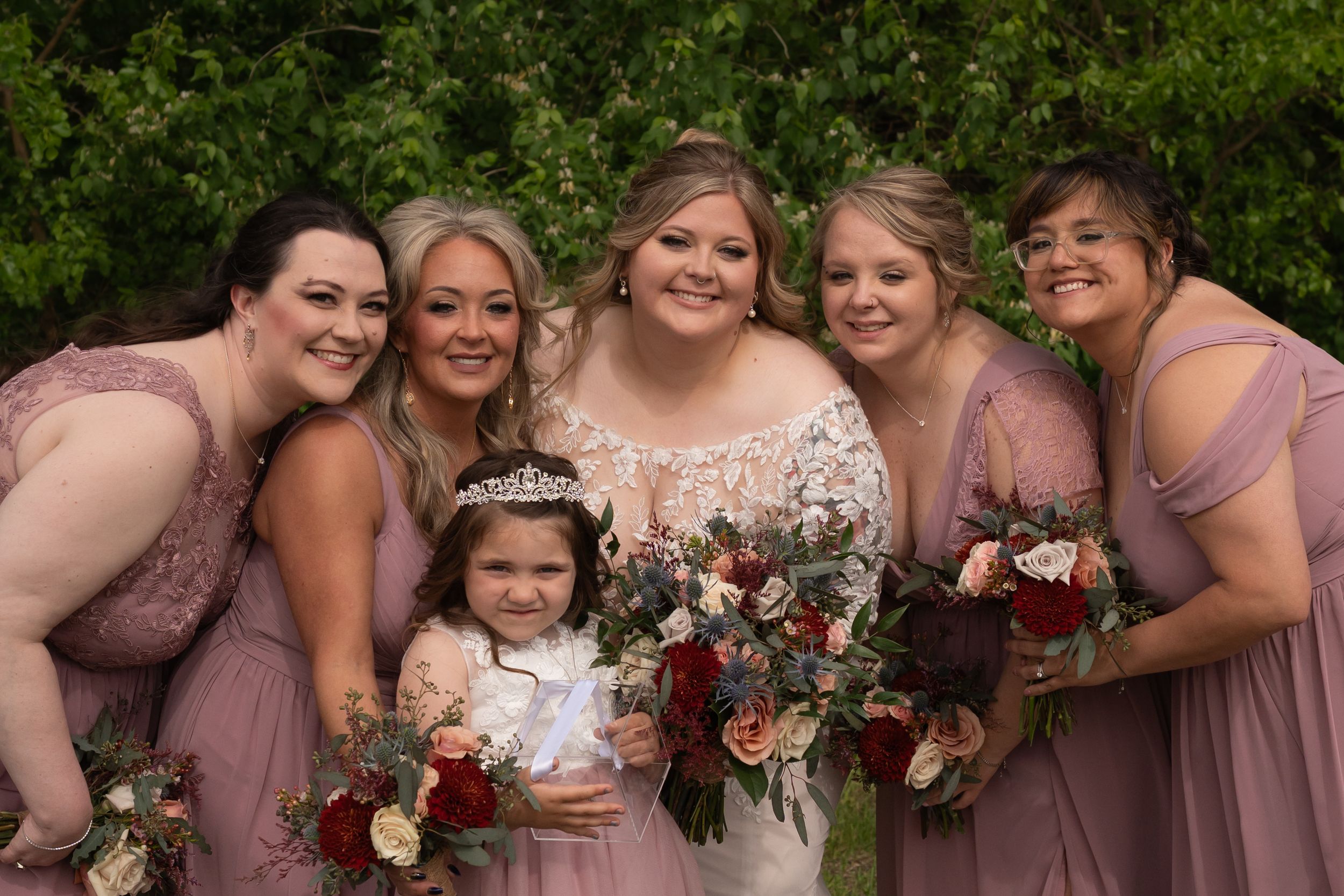 Bride in white lace gown with tiara poses with four bridesmaids in dusty rose dresses and flower girl holding bouquets.
