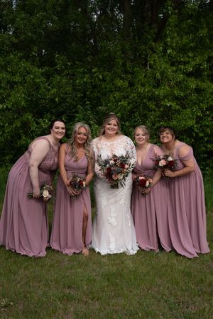 Bride in lace gown poses with four bridesmaids in mauve dresses holding bouquets in front of trees.