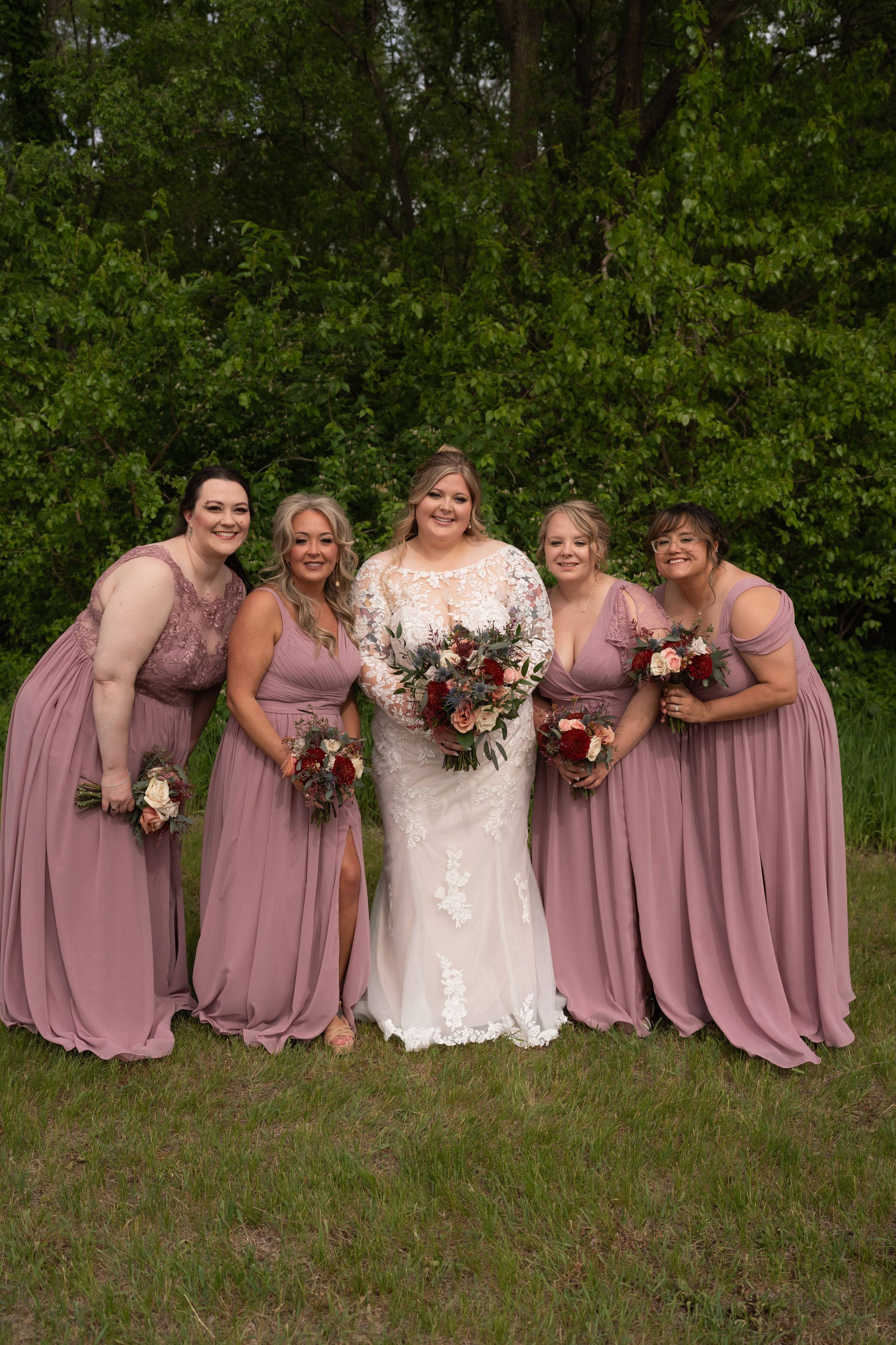 Bride in lace gown poses with four bridesmaids in dusty rose dresses holding bouquets outdoors.