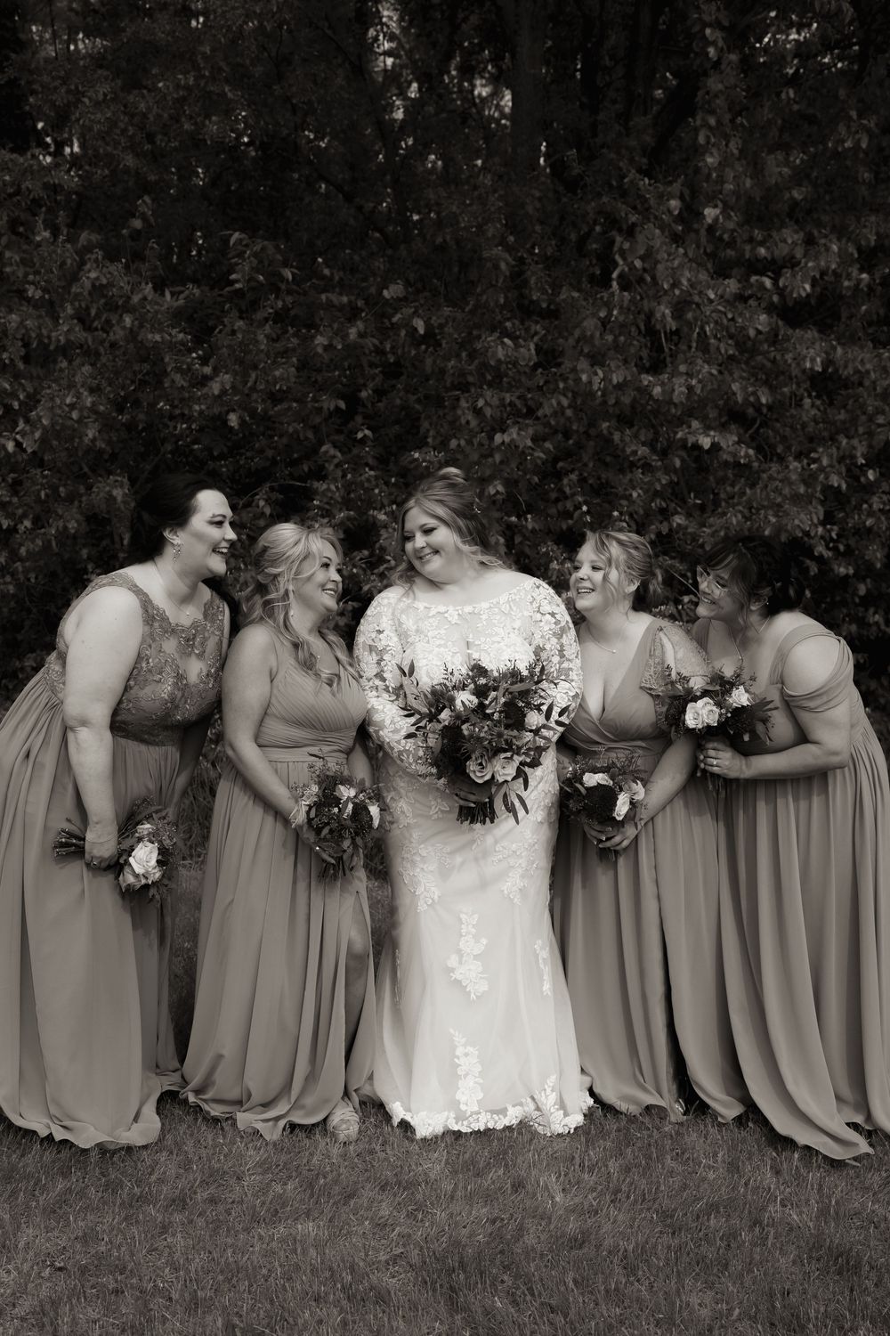 Black and white photo of a bride holding a bouquet surrounded by four bridesmaids in matching gowns outdoors.
