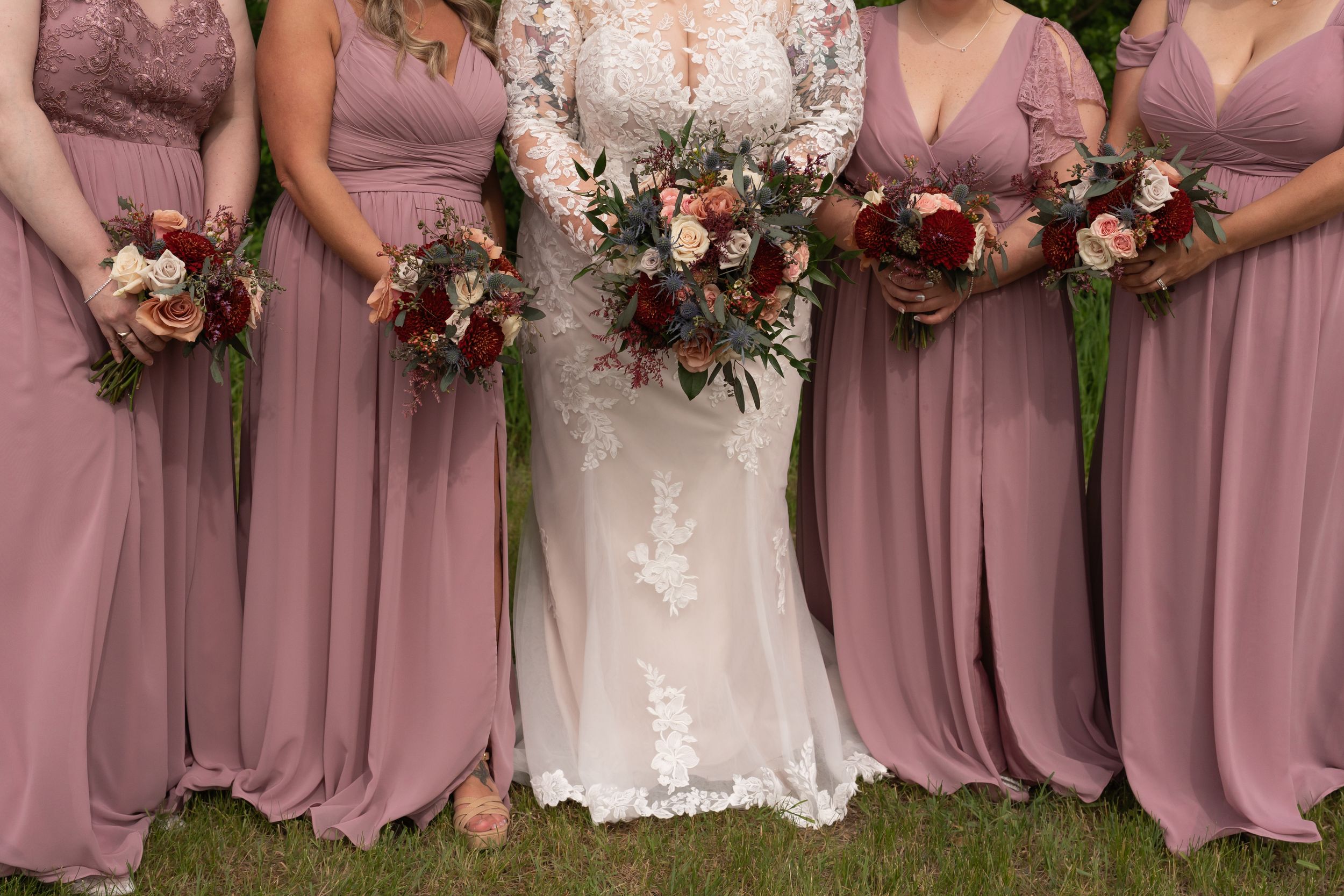 Bride in lace wedding gown stands with bridesmaids in dusty rose gowns holding burgundy and blush floral bouquets.