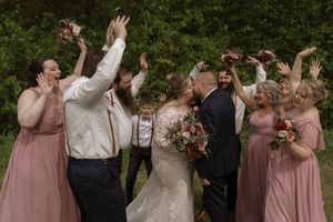 Wedding party celebrates as bride and groom kiss outdoors, bridesmaids in dusty rose gowns raising bouquets joyfully.