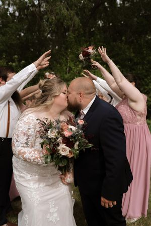 Bride and groom share a kiss under an arch of raised hands formed by their wedding party outdoors.