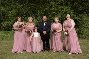 Groom in navy suit poses with four bridesmaids in dusty rose gowns and flower girl outdoors.