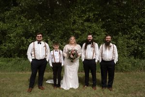 Bride in lace gown poses with groomsmen wearing suspenders and bow ties outdoors.