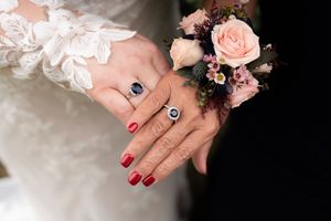 Two hands with sapphire rings and red nails, one wearing a floral corsage, resting on a white lace wedding dress.